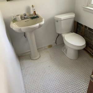 Small bathroom with pedestal sink and toilet, showing missing wall section and exposed subfloor damage.