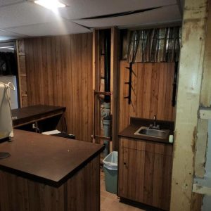 Basement kitchenette with wood cabinets, small sink, and exposed pipes prior to remodeling