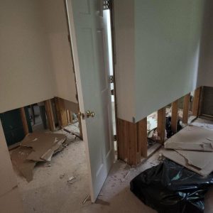 Interior room during water damage restoration with exposed wall studs, removed drywall, debris on the floor, and a door left open.