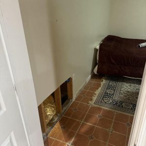 Cut-out section of interior wall exposing insulation after water damage, with tiled floor and furniture nearby.