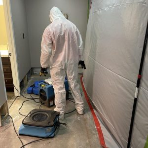 Restoration technician in full protective suit operates air mover and dehumidifier inside a sealed containment area during water damage drying and remediation.