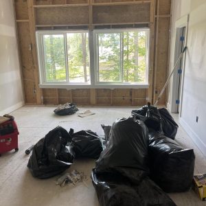 Room under renovation with exposed wall framing, large windows, construction debris, and black trash bags collected after demolition and cleanup work.