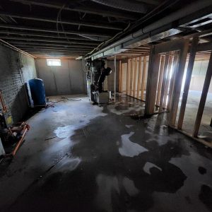 Basement interior with exposed framing and concrete floor showing standing water and moisture damage during flood restoration and cleanup work.
