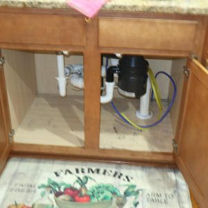 Under-sink cabinet with plumbing pipes, garbage disposal, and hoses visible.