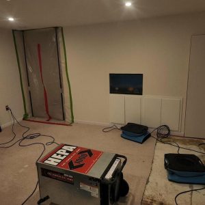 Basement room under restoration with plastic containment barriers, dehumidifiers, air movers, and exposed subfloor during water damage cleanup.