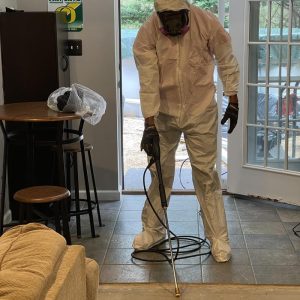 Restoration technician in full protective suit applying antimicrobial treatment to tiled floor during mold remediation and water damage cleanup.