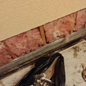 Exposed wall insulation and framing after drywall removal, showing moisture damage and debris along the base of the wall during restoration work.