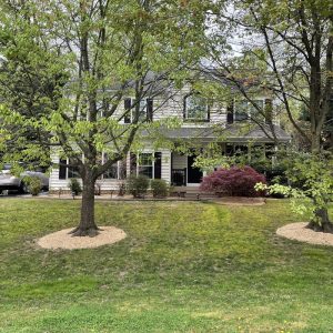 Front exterior of a two-story suburban home surrounded by mature trees and landscaped lawn, photographed during a property inspection or restoration project.