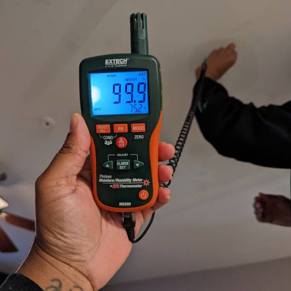 Technician holding a digital moisture meter showing high humidity and moisture levels while inspecting a ceiling during water damage assessment.
